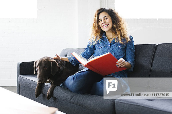 Woman sitting with dog on sofa in living room and reading book
