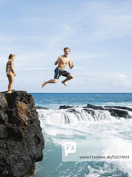 Mädchen (6-7) steht auf einer Klippe und ein Mann springt ins Meer