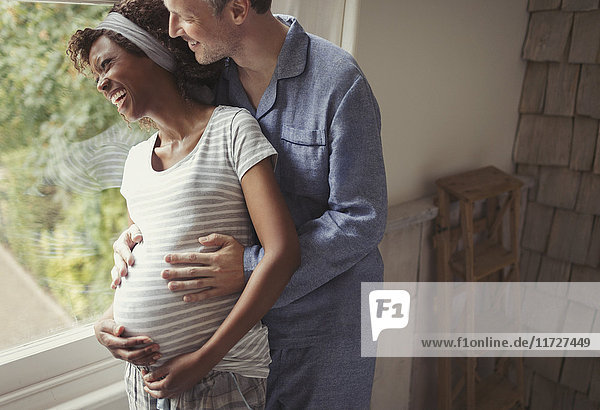 Smiling pregnant couple in pajamas hugging at window
