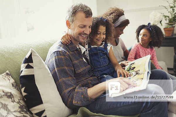 Smiling multi-ethnic father reading book with daughter on sofa