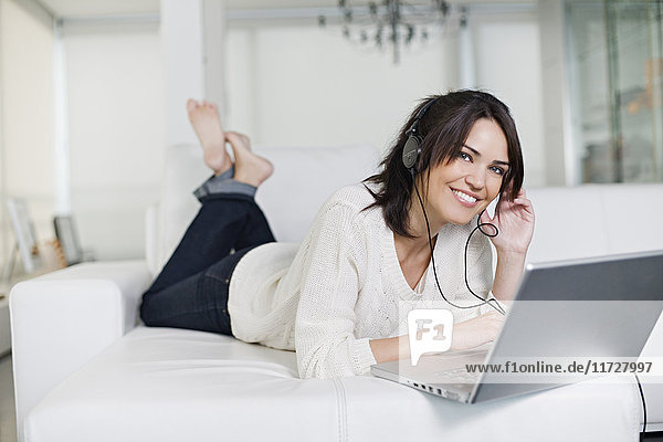 Beautiful brunette woman on a couch with computer and headphones smiling at camera