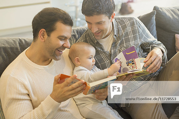Male gay parents reading book to baby son on sofa