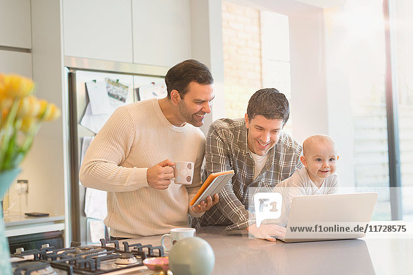 Männliche schwule Eltern mit Baby-Sohn mit digitalem Tablett und Laptop in der Küche