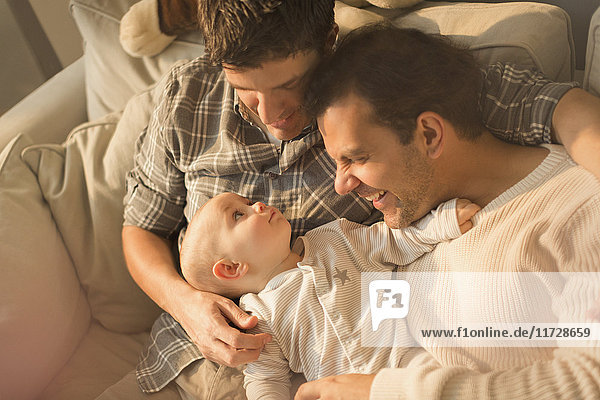 View from above male gay parents cuddling with cute baby son on sofa