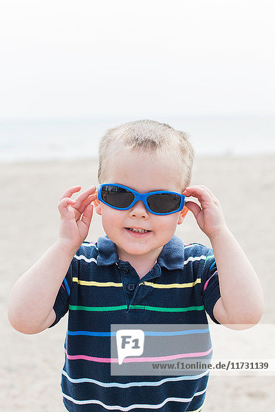 Portrait of male toddler putting on blue sunglasses at beach