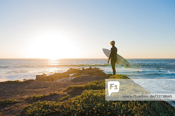 Junger männlicher Surfer schaut bei Sonnenaufgang von der Klippe aufs Meer hinaus