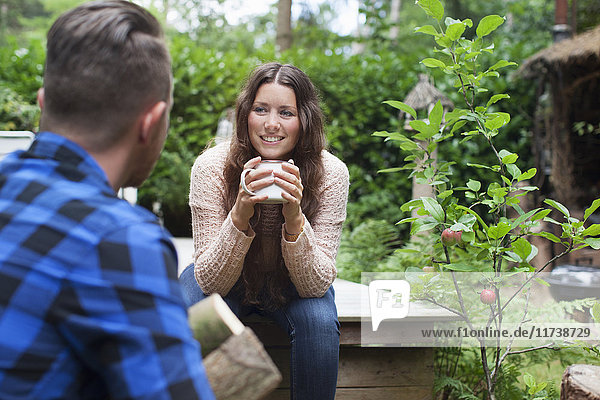 Junge Frau trinkt Kaffee auf der Veranda eines Chalets  während ihr Freund Baumstämme trägt