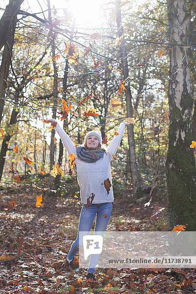 Frontalansicht eines Teenager-Mädchens in voller Länge mit erhobenen Waldarmen  das Herbstblätter wirft und lächelnd aufschaut