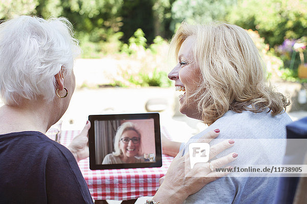 Zwei Frauen sitzen im Freien  halten ein digitales Tablett in der Hand und führen ein Videogespräch mit einem Freund
