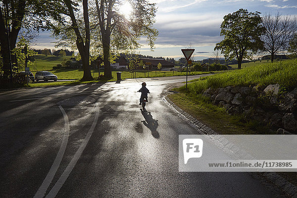 Hintere Silhouettenansicht eines Jungen  der auf einer Landstraße Fahrrad fährt