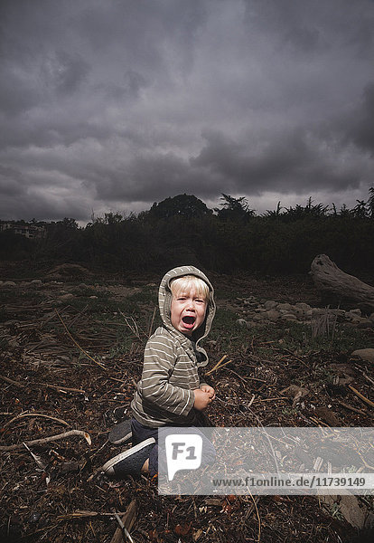 Young boy kneeling on ground crying against stormy sky