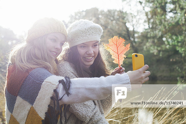 Teenager-Mädchen in Strickwaren am Fluss halten Herbstblatt und benutzen Smartphone  um sich selbst zum Lächeln zu bringen