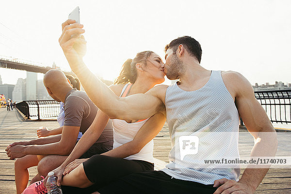 Junger Erwachsener  laufendes Paar küsst sich für Smartphone-Selfie am Flussufer  New York  USA