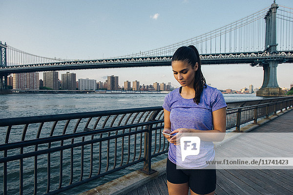 Young female runner reading smartphone in front of Manhattan bridge  New York  USA
