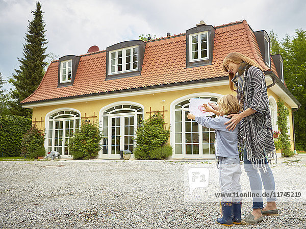Junge zeigt Buntstiftzeichnung einer mittelgroßen erwachsenen Frau  vor dem Haus stehend