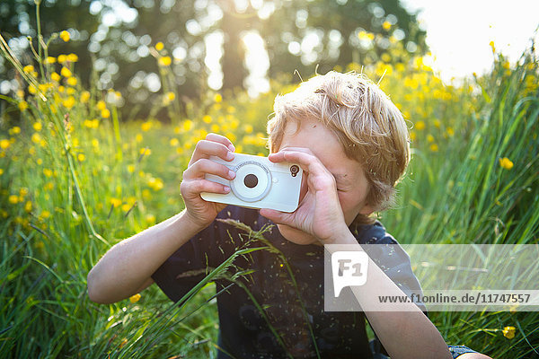 Junge sitzt im hohen Gras und fotografiert mit seinem Smartphone