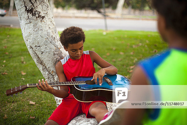 Zwei Brüder sitzen auf einem Ast im Park und spielen akustische Gitarre