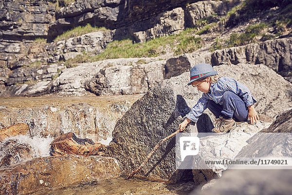 Junge  auf einem Felsen sitzend  mit einem Stock im Schlamm stoßend