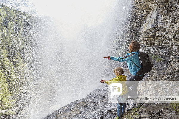Mutter und Sohn  unter einem Wasserfall stehend  strecken die Hände aus  um das Wasser zu spüren  Rückansicht