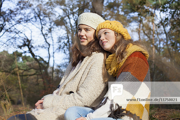 Teenage girls wearing knitwear sitting side by side in forest looking away smiling