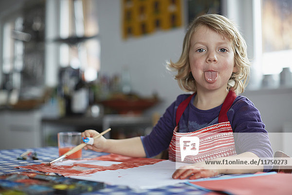 Portrait of young boy sitting at table  holding paintbrush  poking tongue out