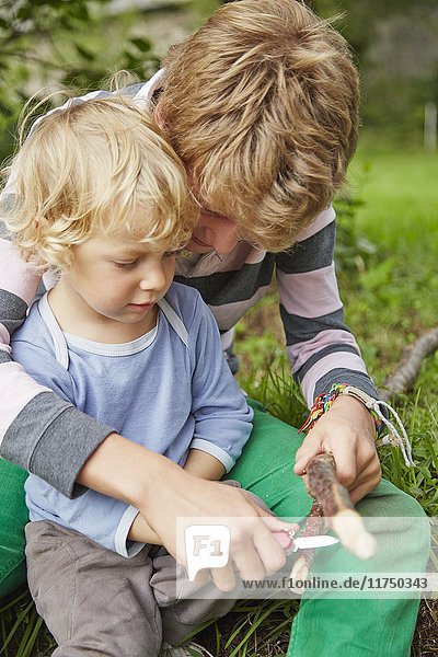 Junge sitzt mit seinem jüngeren Bruder mit einem Taschenmesser auf einem Zweig im Garten