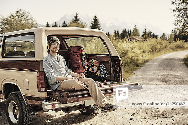 Junger Mann sitzt hinten auf einem Pickup  schaut in die Kamera und lächelt