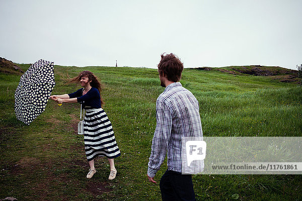 Paar steht an einem windigen Tag im Feld  der Schirm der jungen Frau gerät in den Wind