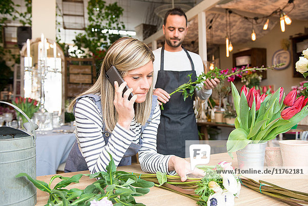 Floristin spricht am Smartphone  ein männlicher Kollege bereitet hinter ihr die Blumenbestellung vor