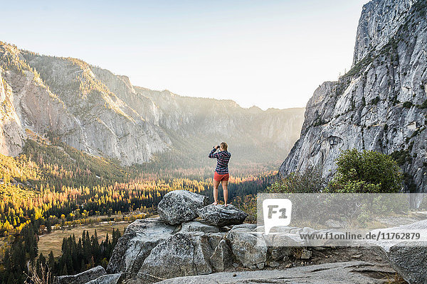 Frau auf Felsbrocken  die durch ein Fernglas auf den Talwald schaut  Yosemite National Park  Kalifornien  USA