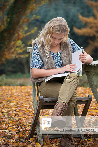 Mädchen sitzt auf Terrassenstuhl und liest Buch im Herbstgarten