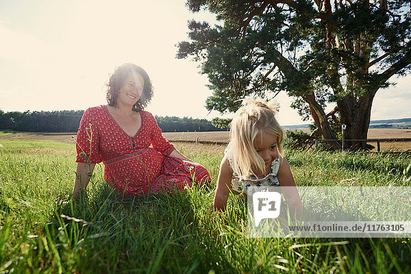 Schwangere Frau sitzt auf dem Feld mit einem Kleinkind  dessen Tochter im Gras krabbelt