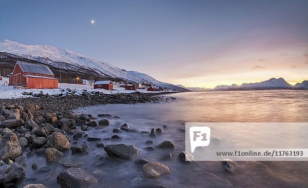 Panorama of typical wood huts called Rorbu framed by fiery sky at sunset and frozen sea Djupvik Lyngen Alps Troms Norway Scandinavia Europe