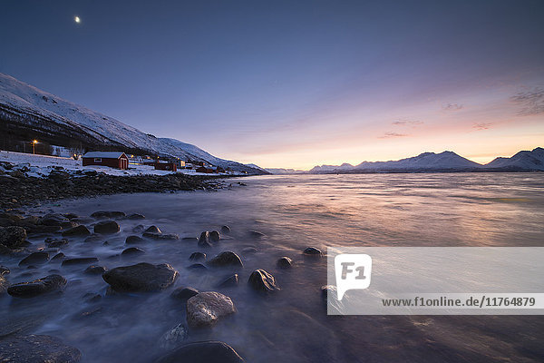 Feuriger Himmel bei Sonnenuntergang auf typischem Rorbu  eingerahmt von schneebedeckten Gipfeln und gefrorenem Meer  Djupvik  Lyngen Alps  Troms  Norwegen  Skandinavien  Europa