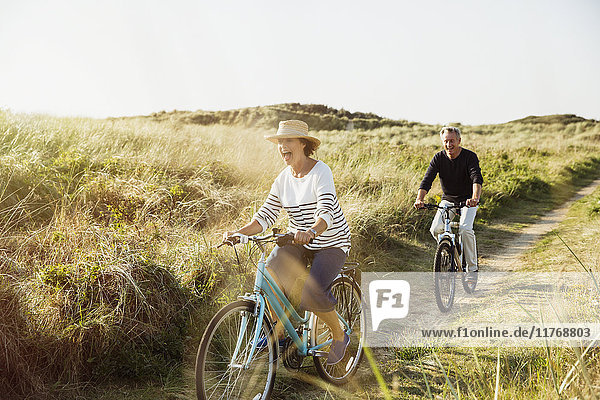 Playful mature couple riding bicycles on sunny beach grass path
