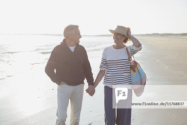 Lächelndes reifes Paar hält sich an den Händen und geht am sonnigen Strand spazieren.