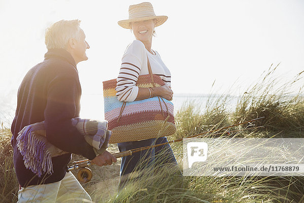 Smiling mature couple with fishing rod walking in sunny beach grass