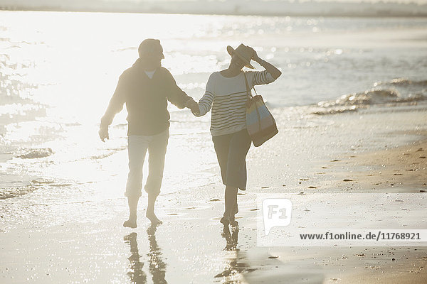 Ein reifes Paar hält sich an den Händen und geht am sonnigen Strand spazieren.