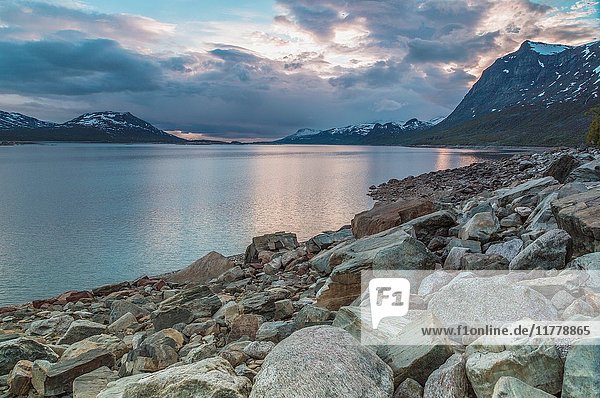 Sun reflecting in water  mountains with snow in background and a rocky beach in foreground  Stora sjöfallets national park  Gällivare  Swedish Lapland  Sweden.