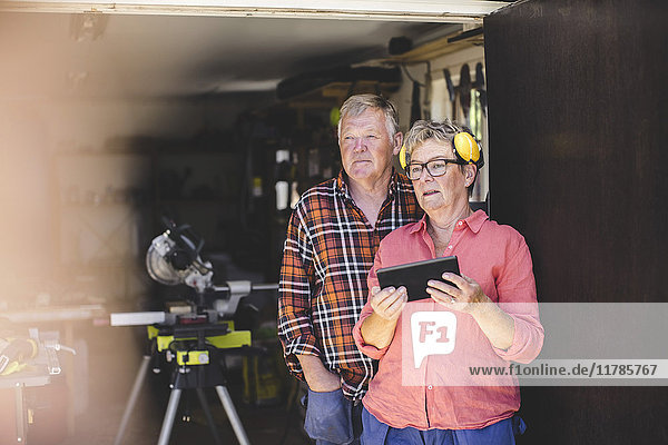 Nachdenkliches Seniorenpaar mit digitalem Tablett im Stehen vor der Werkstatttür
