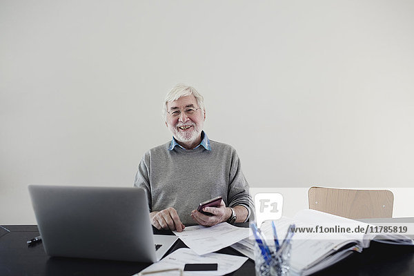 Portrait of smiling senior man with laptop and bills sitting against white wall