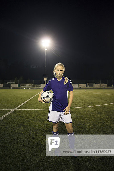 Porträt eines selbstbewussten Mädchens  das nachts den Ball auf dem Fußballfeld gegen den Himmel hält.
