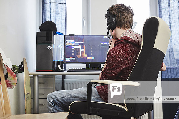 Teenager Junge mit Kopfhörer am Computer  während er zu Hause auf dem Stuhl sitzt.