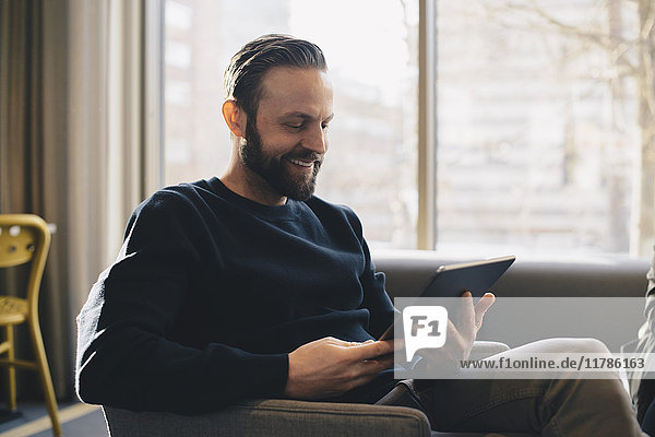 Happy businessman using digital tablet while sitting on sofa in office lobby