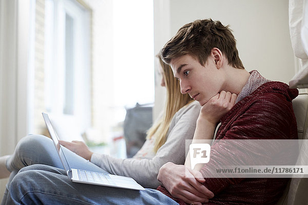 Teenager Junge beobachtet Laptop  während er von seiner Schwester im Wohnzimmer zu Hause sitzt.