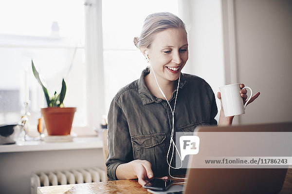 Happy woman listening to headphones while holding coffee cup at home