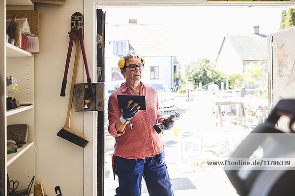 Seniorin mit Bohrer und digitalem Tablett im Stehen am Eingang der Werkstatt