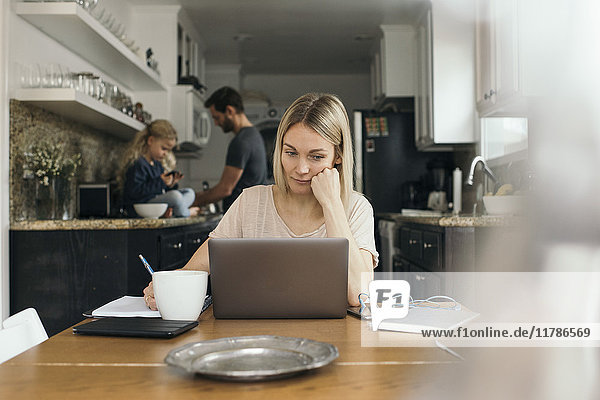 Mittlere erwachsene Frau mit Laptop am Tisch mit Familie in der Küche zu Hause