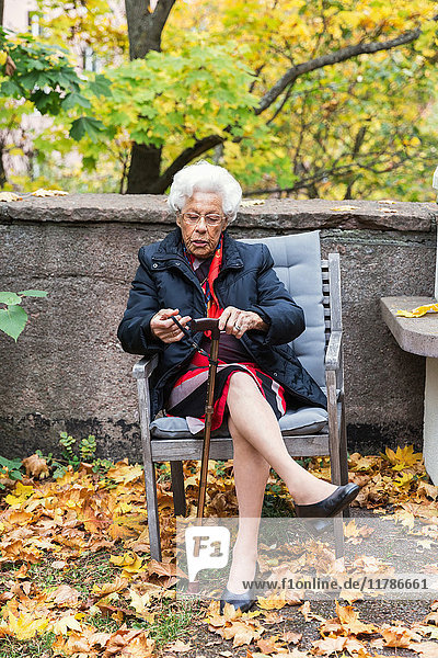 Volle Länge der älteren Frau  die auf einem Stuhl im Park sitzt und einen Stock hält.