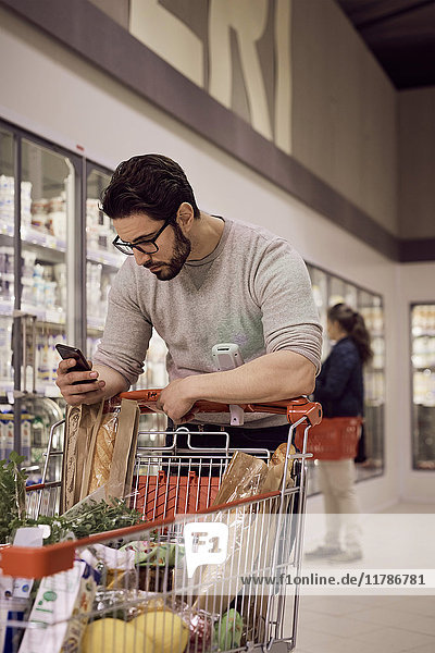 Man Textnachrichten  während man sich auf den Einkaufswagen im Kühlregal im Supermarkt stützt.